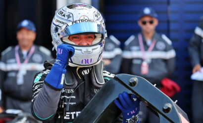 Formula 1 driver in a Petronas helmet and blue gloves holds a car component while team members watch in the background.