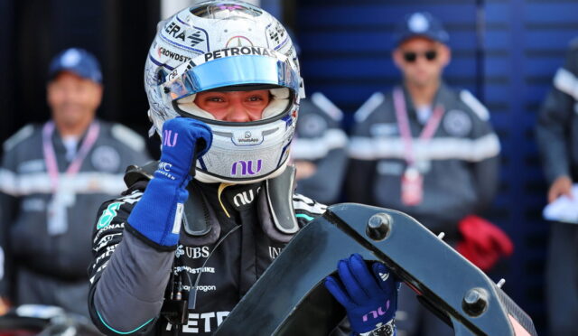 Formula 1 driver in a Petronas helmet and blue gloves holds a car component while team members watch in the background.