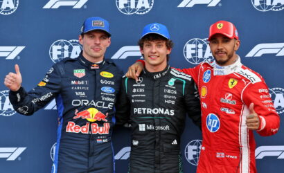Three Formula 1 drivers pose on a podium in racing suits (Red Bull, Mercedes Petronas, Ferrari) with thumbs up in front of FIA backdrop.