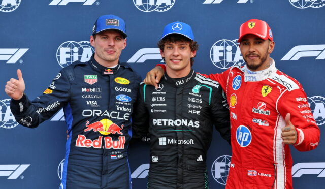 Three Formula 1 drivers pose on a podium in racing suits (Red Bull, Mercedes Petronas, Ferrari) with thumbs up in front of FIA backdrop.