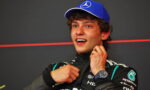 Young race car driver in a black Petronas racing suit and a blue Mercedes cap, smiling and adjusting his collar after a race.
