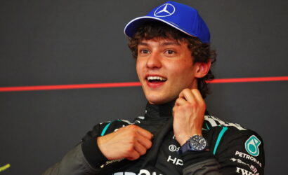 Young race car driver in a black Petronas racing suit and a blue Mercedes cap, smiling and adjusting his collar after a race.