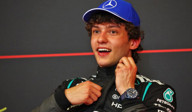 Young race car driver in a black Petronas racing suit and a blue Mercedes cap, smiling and adjusting his collar after a race.