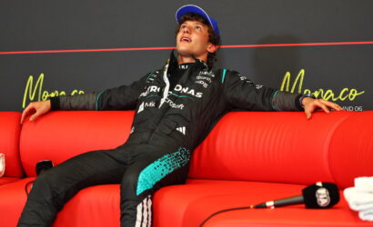 Young race car driver in a black teal racing suit lounges on a bright red couch, arms stretched along the back, looking upward.