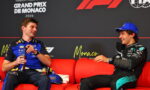 Two Formula 1 drivers sit on a red couch during a Monaco Grand Prix interview, smiling at each other against a branded backdrop.
