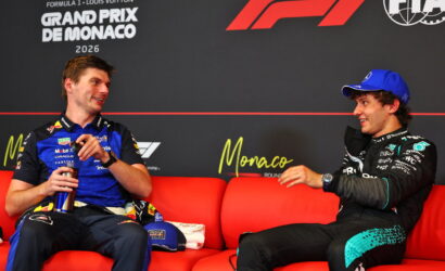 Two Formula 1 drivers sit on a red couch during a Monaco Grand Prix interview, smiling at each other against a branded backdrop.
