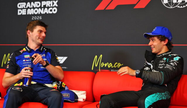 Two Formula 1 drivers sit on a red couch during a Monaco Grand Prix interview, smiling at each other against a branded backdrop.