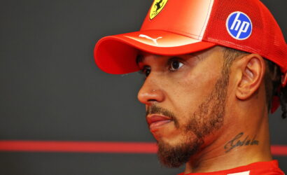 Man in profile wearing a red Ferrari cap and red shirt, neck tattoo visible on his left side, against a dark background.