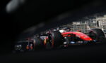 Red Ferrari Formula 1 car racing along a city street circuit, seen through a foreground obstruction with blurred edges and spectators in the background