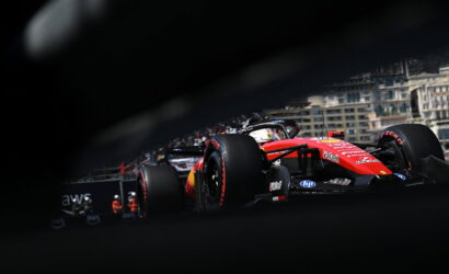 Red Ferrari Formula 1 car racing along a city street circuit, seen through a foreground obstruction with blurred edges and spectators in the background
