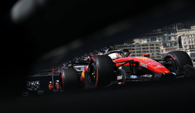 Red Ferrari Formula 1 car racing along a city street circuit, seen through a foreground obstruction with blurred edges and spectators in the background