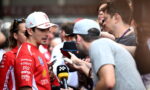 Race car driver in a red suit answers questions from reporters holding microphones and phones.