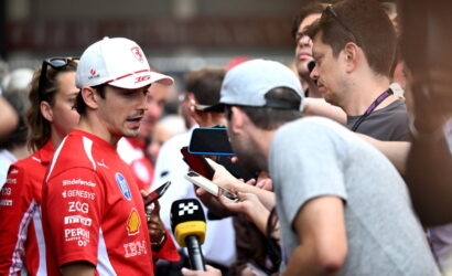 Race car driver in a red suit answers questions from reporters holding microphones and phones.