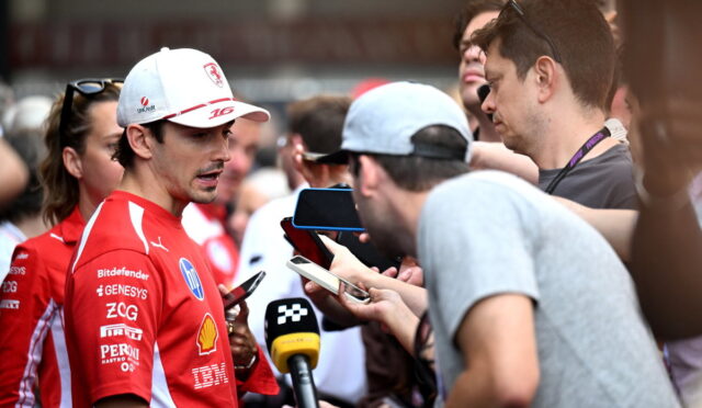 Race car driver in a red suit answers questions from reporters holding microphones and phones.