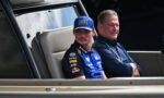 Racing driver wearing a blue suit and cap sits in a vehicle's cabin beside another man in a dark polo shirt, both looking ahead.