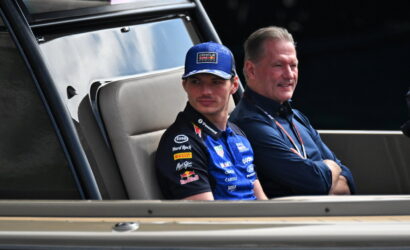 Racing driver wearing a blue suit and cap sits in a vehicle's cabin beside another man in a dark polo shirt, both looking ahead.