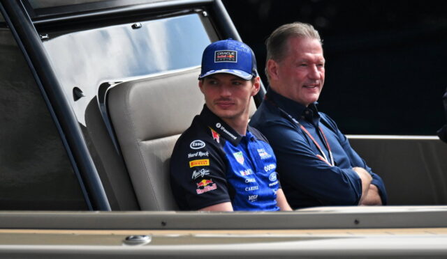 Racing driver wearing a blue suit and cap sits in a vehicle's cabin beside another man in a dark polo shirt, both looking ahead.