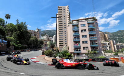 Formula 1 cars race along a winding urban street circuit past high-rise buildings and balconies of spectators.