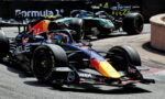 Red Bull Racing F1 car leading a tight corner, with a Mercedes car close behind on a race track barrier backdrop behind them