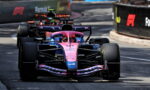 Pink Formula 1 car leading a race on a street circuit, close view with a second car close behind, barriers on sides.