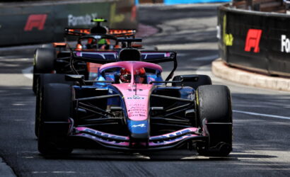 Pink Formula 1 car leading a race on a street circuit, close view with a second car close behind, barriers on sides.