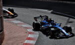 Blue Formula 1 car with Barclays livery rounds a street-circuit kerb, a yellow car trailing in the background on the left curb.