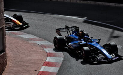 Blue Formula 1 car with Barclays livery rounds a street-circuit kerb, a yellow car trailing in the background on the left curb.