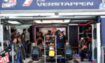 Inside a Red Bull Racing garage, a Formula 1 car is elevated on a stand while crew members work around it under a large banner with a driver’s name and the Red Bull logo.
