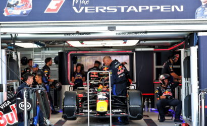 Inside a Red Bull Racing garage, a Formula 1 car is elevated on a stand while crew members work around it under a large banner with a driver’s name and the Red Bull logo.