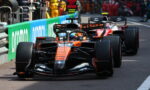 Formula 1 cars in a pit lane, orange car leading followed by red and black cars with crew in the background.