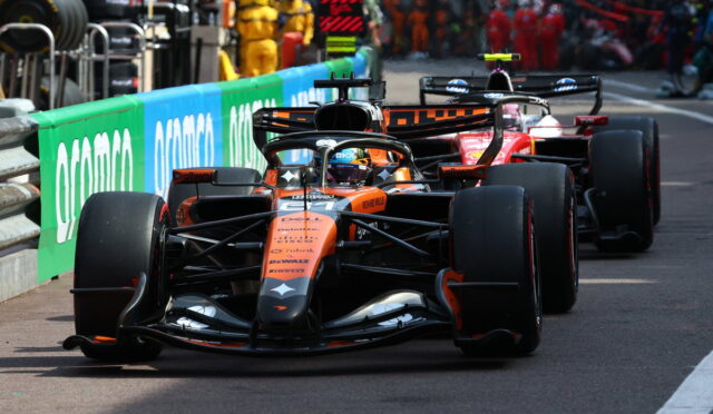 Formula 1 cars in a pit lane, orange car leading followed by red and black cars with crew in the background.