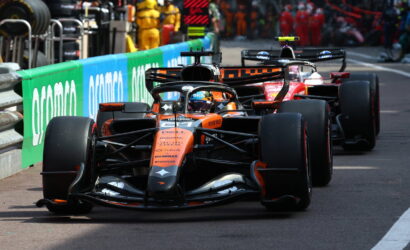 Orange McLaren Formula 1 car leads a pack on the pit straight, with crew and barriers in the background.