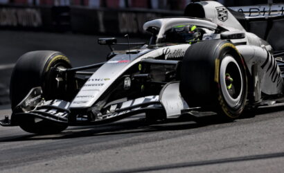 White Formula 1 car corners on a race track, driver in helmet visible inside the cockpit, Cadillac branding on the nose and side.