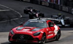 Red Mercedes AMG safety car leading a pack of Formula 1 cars on a race track, sponsor logos visible on the bodywork and hood.