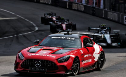 Red Mercedes AMG safety car leading a pack of Formula 1 cars on a race track, sponsor logos visible on the bodywork and hood.