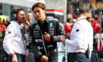 Young Formula 1 driver in a black Petronas racing suit being interviewed by two engineers in white shirts with headsets in a busy pit lane.