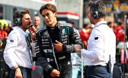 Young Formula 1 driver in a black Petronas racing suit being interviewed by two engineers in white shirts with headsets in a busy pit lane.