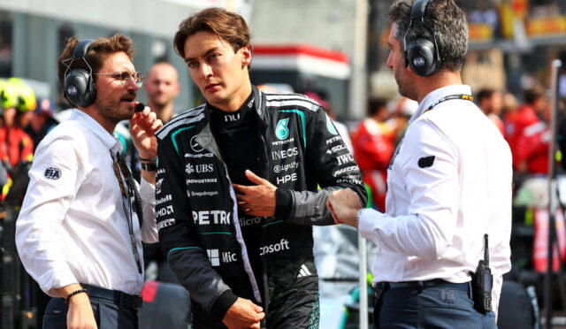 Young Formula 1 driver in a black Petronas racing suit being interviewed by two engineers in white shirts with headsets in a busy pit lane.