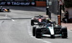 Formula 1 cars race on a city street circuit; the Mercedes car leads with a second red car close behind, barriers and a TAG Heuer banner in view.