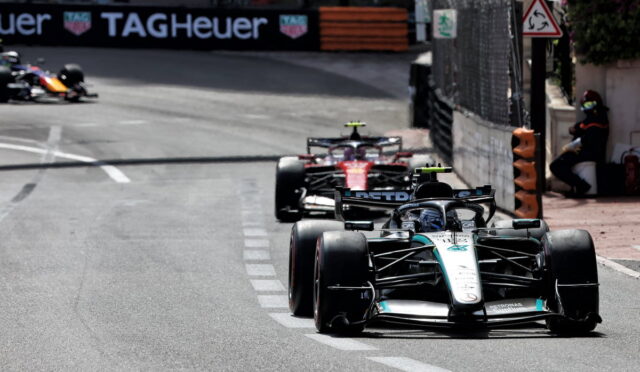 Formula 1 cars race on a city street circuit; the Mercedes car leads with a second red car close behind, barriers and a TAG Heuer banner in view.