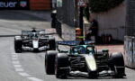Two Formula 1 cars racing on a city street circuit; a teal-green car leads the silver car in the background with barriers and signage nearby.