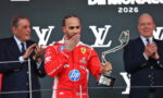 Ferrari driver in a red racing suit on a podium kisses a tall abstract trophy as two suited officials applaud behind him, Monaco 2026 backdrop visible.