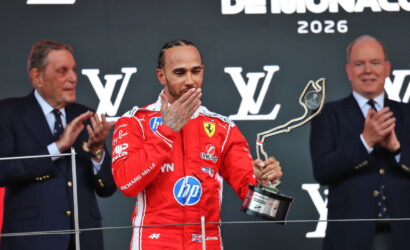 Ferrari driver in a red racing suit on a podium kisses a tall abstract trophy as two suited officials applaud behind him, Monaco 2026 backdrop visible.