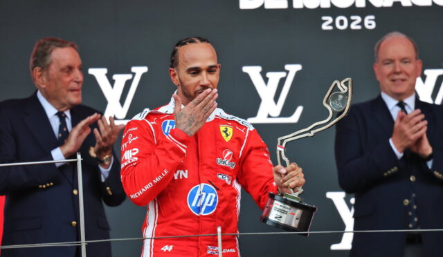 Ferrari driver in a red racing suit on a podium kisses a tall abstract trophy as two suited officials applaud behind him, Monaco 2026 backdrop visible.