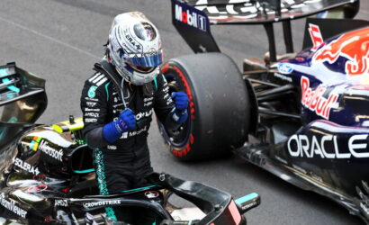 Mercedes-Petronas Formula 1 driver in a black racing suit and silver helmet raises fists in celebration beside his car on track, with a Red Bull car in the background behind him.