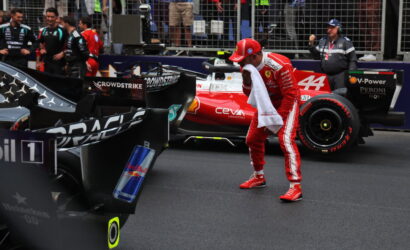 F1 driver in a red racing suit wipes sweat with a towel beside race cars in the pit lane.