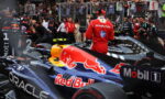 F1 pit lane with a Red Bull racing car in the foreground and a driver in a red suit standing beside it, photographers and spectators behind the fence