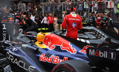 F1 pit lane with a Red Bull racing car in the foreground and a driver in a red suit standing beside it, photographers and spectators behind the fence
