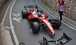 Red Formula 1 race car crashed on a city street, with its front wing detached and debris nearby, while a marshal signals nearby against a stone wall backdrop.