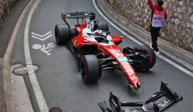 Red Formula 1 race car crashed on a city street, with its front wing detached and debris nearby, while a marshal signals nearby against a stone wall backdrop.